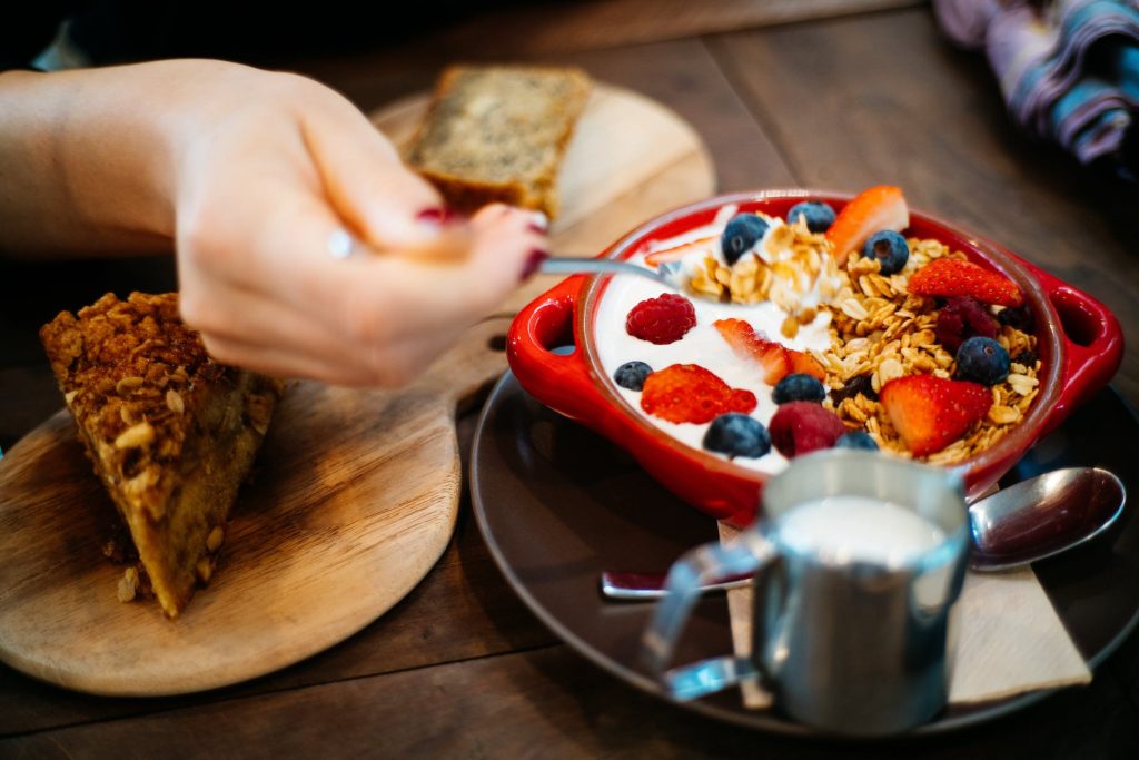 person holding spoon and round red ceramic bowl with pastries